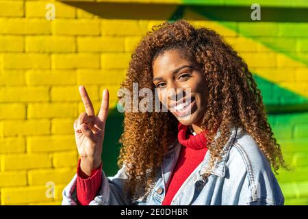 Jeune femme noire optimiste millénaire avec des cheveux bouclés dans un style élégant des vêtements montrant des gestes avec les doigts et souriant tout en regardant caméra contre y Banque D'Images