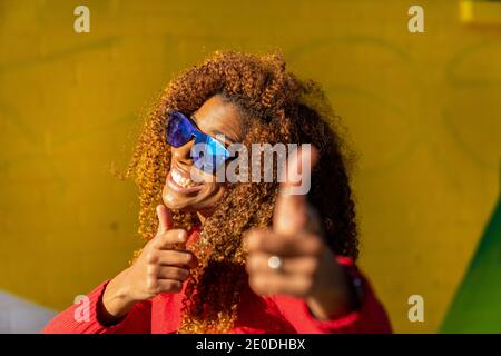 Jeune femme afro-américaine gaie avec des cheveux bouclés dans une ambiance décontractée vêtements et lunettes de soleil souriant et pointant vers l'appareil photo contre la lumière vive mur coloré Banque D'Images