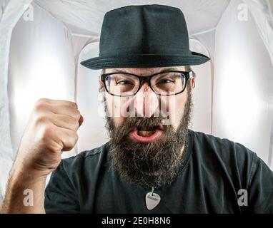 Un homme barbu et furieux, qui ressemble à un artiste ou à un musicien avec des lunettes noires et un chapeau montrant le poing Banque D'Images