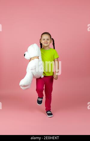 Une fille à queue de cochon pleine de contentement dans un t-shirt vert clair et un pantalon rouge marche avec un ours en peluche blanc isolé sur fond rose Banque D'Images