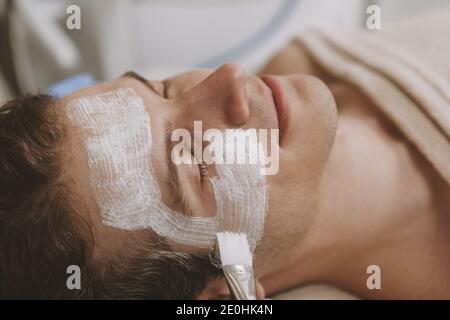 Petit gros plan d'un beau homme détendu qui obtient un masque facial par un cosmétologue professionnel au salon de beauté. Client masculin attrayant se reposant au spa cent Banque D'Images