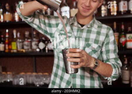 Coupe courte de barman professionnel joyeux préparant un cocktail pour un client, avec un moule à shaker en acier Banque D'Images