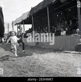 Dans les années 1960, deux jeunes enfants, historiques, se lançant dans une course amusante sur l'herbe sous la surveillance d'une foule de spectateurs assis dans une tribune abritée en bois à un Highland Games, Highlands, Écosse, Royaume-Uni. Banque D'Images