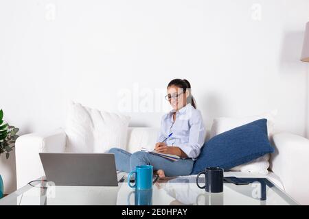Jeune femme adulte prenant des notes, avec son ordinateur portable sur le canapé dans le salon à la maison. Avec espace de copie. Banque D'Images