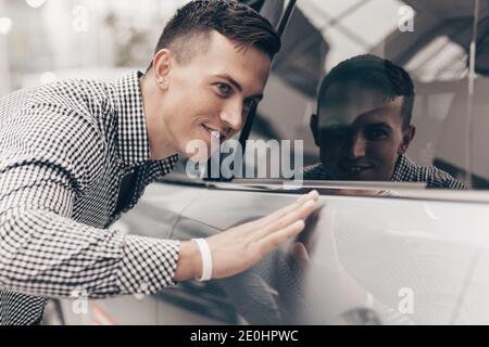 Gros plan d'un bel homme heureux touchant doucement la nouvelle voiture à la concession. Un client masculin attractif examine les voitures en vente au salon automobile showroo Banque D'Images