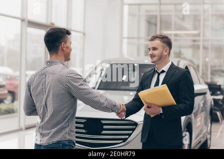 Vendeur de voitures professionnel souriant, se serrant la main avec son client masculin devant une nouvelle automobile. Homme achetant une nouvelle voiture au salon, en serrant les mains W Banque D'Images