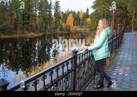 Une femme blonde dans une veste se tient sur le remblai de la rivière dans la forêt d'automne parmi les grands pins. Passer du temps dans la nature. Banque D'Images