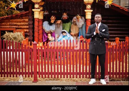 Portrait du jeune et beau homme d'affaires afro-américain en costume séjour et priez contre la crèche de Noël et la scène de la nativité. Banque D'Images