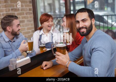 Joyeux barbu homme souriant à la caméra tout en buvant de la bière avec ses amis au pub Banque D'Images