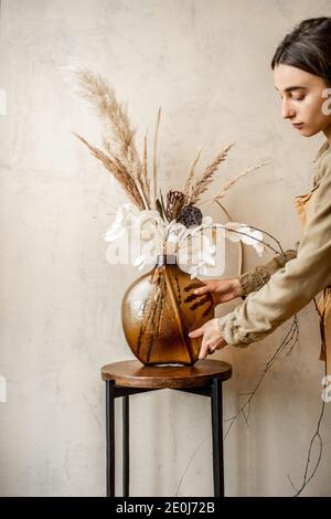 Femme décorant la maison avec une composition de fleurs séchées et herbes dans un vase en verre sur un fond de mur beige Banque D'Images