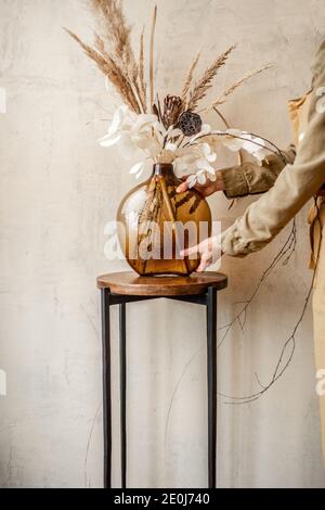 Femme décorant la maison avec une composition de fleurs séchées et herbes dans un vase en verre sur un fond de mur beige Banque D'Images