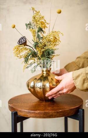 Maison de décodage femelle avec composition de fleurs fraîches et d'herbes dans un vase doré sur un fond de mur beige. Bouquet d'acacia, dealbata, mimosa, nelumbo et Craspedia Banque D'Images
