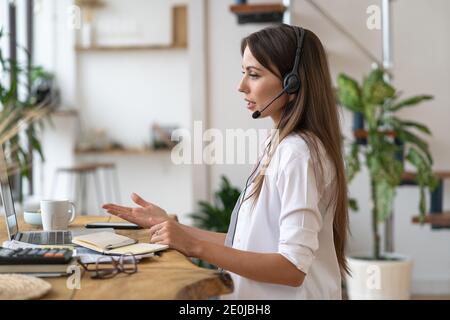 Vue latérale d'une femme caucasienne pigiste dans des écouteurs assis à la table, parlant dans le chat vidéo, travaillant à distance depuis le bureau à domicile. Travail à distance. Banque D'Images