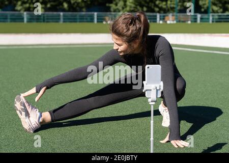 Femme en sport vlogger assis sur gazon artificiel en extérieur, montrant et démontrant des exercices pour son blog en ligne, des enregistrements sur smartphone caméra Banque D'Images