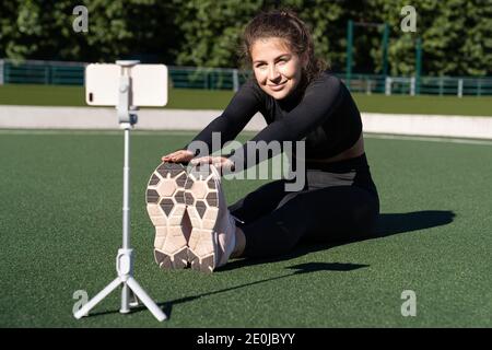 Femme souriante vlogger dans les vêtements de sport assis sur le gazon artificiel en extérieur, démontrant des exercices pour son blog en ligne, des enregistrements sur smartphone caméra sur t Banque D'Images