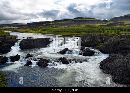 Chute d'eau de Glanni sur la rivière Norðurá dans l'ouest de l'Islande. L'eau coule au-dessus de la roche de lave noire et les montagnes s'élèvent des rives de la rivière. Banque D'Images