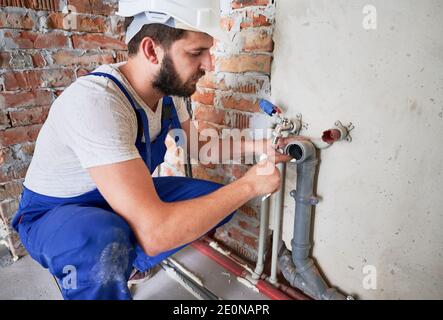 Jeune plombier, portant un uniforme bleu et un casque blanc travaillant avec le produit d'étanchéité de la pipe d'égout dans la cuisine ou dans la salle de bains dans l'appartement inachevé. Installation du concept de système de plomberie Banque D'Images