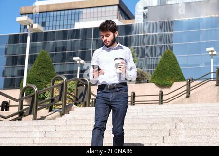Jeune homme d'affaires avec une tasse de café dans sa main et en vérifiant son smartphone marchant dans les escaliers. Homme d'affaires dans une rue urbaine d'une ville. Banque D'Images