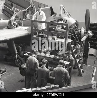 Années 1950, historique, portant un manteau blanc, un ingénieur senior qui supervise un groupe de jeunes hommes stagiaires ou apprentis en génie de l'aviation, travaillant sur le moteur d'un avion de formation avec une hélice, Angleterre, Royaume-Uni. Banque D'Images