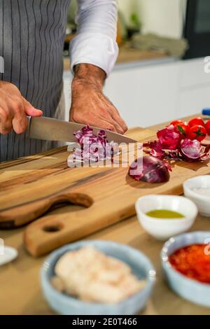 Le chef coupe des oignons dans la cuisine sur une planche de bois. Il y a des légumes, des épices et de petits bols de cuisine autour de la surface de travail. Banque D'Images