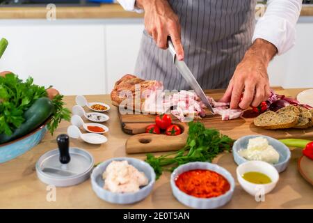 Le chef coupe du bacon dans la cuisine sur une planche à bois. Il y a des légumes, des tranches de pain, des épices et de petits bols de cuisine autour de la surface de travail. Banque D'Images