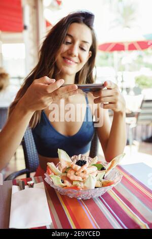 Jeune femme photographiant sa salade avec un smartphone tout en étant assise dans un restaurant Banque D'Images