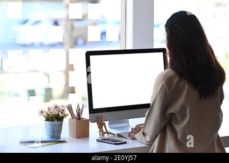 Vue arrière d'une jeune femme travaillant sur un ordinateur dans un bureau moderne. Écran vide pour le montage du produit. Banque D'Images