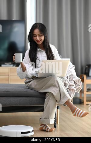 Une femme heureuse assise sur un canapé, travaillant avec un ordinateur portable et buvant du café, tandis que le robot aspire le sol à la maison. Banque D'Images