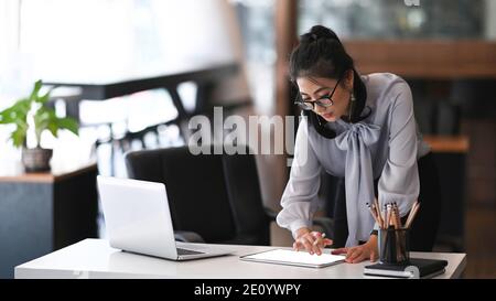 Une jeune femme designer de lunettes de vue travaillant avec un ordinateur portable et une tablette numérique tout en étant debout dans un bureau moderne. Banque D'Images