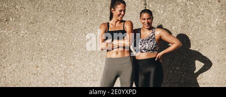 Prise de vue panoramique de deux femelles se détendant après l'entraînement. Les femmes sont amies sur des vêtements de sport debout ensemble et souriantes. Banque D'Images