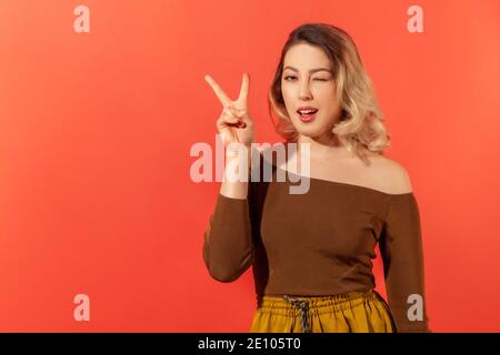 Portrait d'une jeune femme heureuse avec des cheveux blonds dans un chemisier marron montrant le geste de victoire par ses doigts et en se faisant un clin d'œil à l'appareil photo. Goujon intérieur Banque D'Images