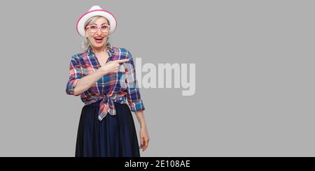 Portrait de femme mature élégant moderne surpris dans un style décontracté avec chapeau et lunettes debout, étonné à la caméra et en montrant au mur. ind Banque D'Images