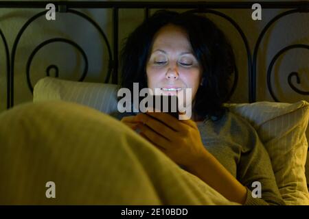Une femme hispanique contentée vérifie ses messages téléphoniques ou regarde les médias comme elle se détend dans le lit la nuit avec un sourire allumé par la lueur de l'écran Banque D'Images
