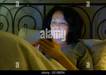 Une femme hispanique contentée vérifie ses messages téléphoniques ou regarde les médias comme elle se détend dans le lit la nuit avec un sourire allumé par la lueur de l'écran Banque D'Images