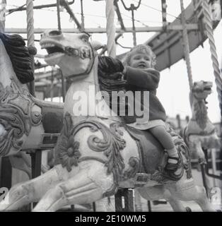 Années 1950, historique, une jeune fille à cheval sur un cheval en bois lors d'une promenade en carrousel dans un parc d'expositions, Angleterre, Royaume-Uni. Un carrousel est un tour d'amusement qui a une plate-forme rotative, également connu comme un joyeux-Go-round. Il est inhabituel de voir un enfant si jeune en solo qu'ils sont normalement accompagnés d'un adulte, mais Hé, c'est les années 1960! Banque D'Images