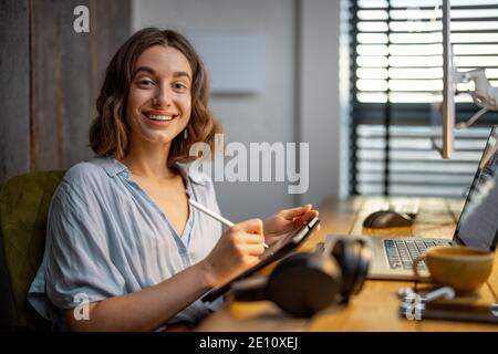 Portrait d'une jeune femme en tant qu'artiste numérique, tirant sur une tablette numérique dans le lieu de travail confortable à la maison Banque D'Images