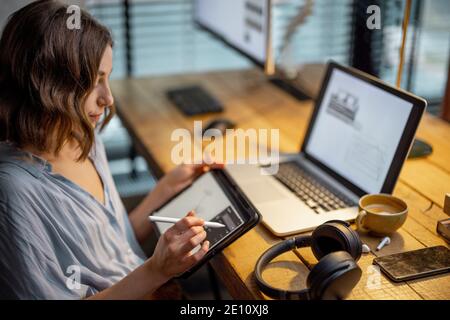 Jeune femme vêtue à la manière décontractée ayant un peu de travail créatif, dessin sur une tablette numérique, assis dans le bureau à la maison confortable et élégant Banque D'Images