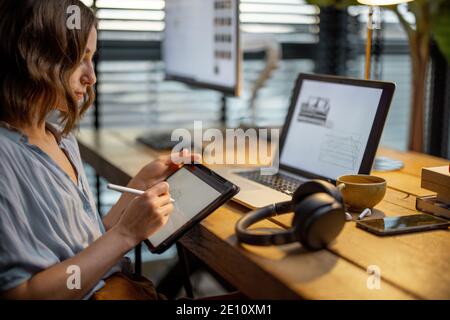 Jeune femme vêtue à la manière décontractée ayant un peu de travail créatif, dessin sur une tablette numérique, assis dans le bureau à la maison confortable et élégant Banque D'Images