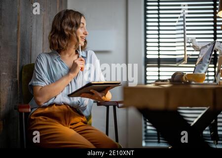 Jeune femme vêtue à la manière décontractée ayant un peu de travail créatif, dessin sur une tablette numérique, assis dans le bureau à la maison confortable et élégant Banque D'Images