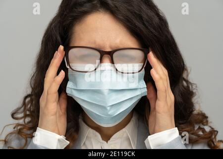 Femme d'affaires avec des lunettes brumeuses de souffle causé par le port masque jetable sur le mur gris studio Banque D'Images