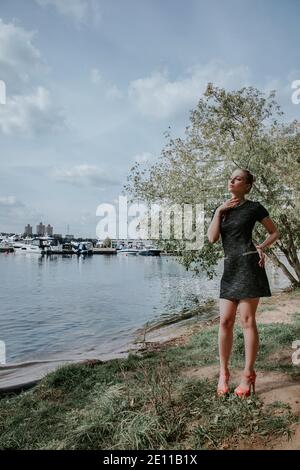 Jolie fille blonde se tient dans le parc près de l'eau avec des bateaux à l'arrière-plan; elle porte une robe courte et élégante sombre et des talons hauts Banque D'Images