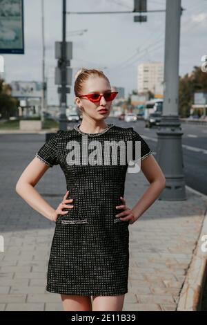 Jolie fille blonde se tient dans une ville; elle porte une robe courte et élégante, des lunettes de soleil rouges et des talons hauts; des voitures passent dans une rue animée Banque D'Images