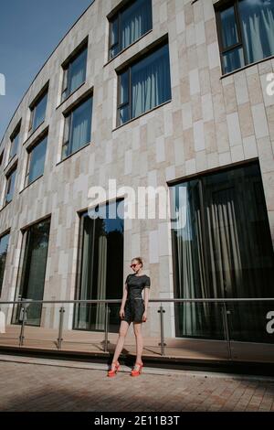 Jolie fille blonde se tient devant un bâtiment urbain moderne; elle porte une robe courte et élégante, des lunettes de soleil rouges et des talons hauts Banque D'Images