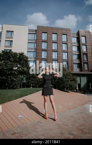 Jolie fille blonde se tient dans un environnement urbain moderne ; elle porte une robe courte et élégante avec des talons hauts ; elle semble tenir ses lunettes de soleil rouges Banque D'Images