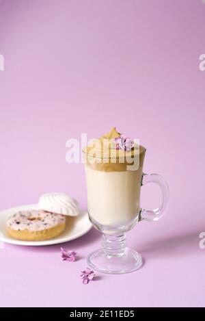 Belle Dalgona buvez un café mousseux dans un mug transparent et des fleurs de lilas sur fond violet. Bonbons sur une assiette: Donat et marshmal tendre Banque D'Images