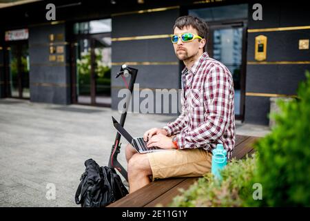 Décontracté et attrayant, le jeune homme de caucasien s'assoit sur un banc à côté de son scooter électrique en utilisant un ordinateur portable pour travailler à distance. Freelance et la technologie moderne font Banque D'Images