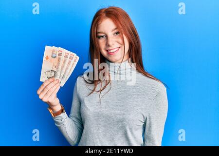 Jeune fille à tête rouge détenant 10 billets de banque au royaume-uni avoir l'air positif et heureux debout et sourire avec une assurance sourire montrant les dents Banque D'Images