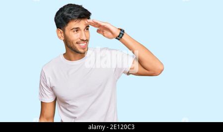 Jeune homme beau portant une chemise blanche décontractée très heureux et souriant regardant loin avec la main sur la tête. Concept de recherche. Banque D'Images