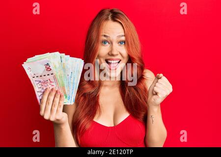 La jeune femme à tête rouge qui détient des billets de banque en dollars de hong kong criait fière, célébrant la victoire et le succès très excitée par les armes levées Banque D'Images