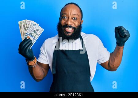 Un jeune homme afro-américain portant un tablier professionnel tenant des billets de banque en dollars américains criant fier, célébrant la victoire et le succès très excité avec Banque D'Images